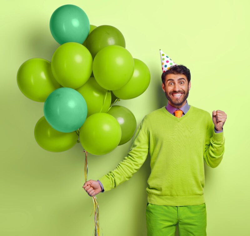 A man holding balloons, wearing a party hat and clenching his fist in delight. Everything is green except for his shirt which is purple and his. tie which is orange. 