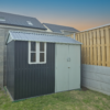 The 10ft x 6ft Steel Cottage shed in a garden. There is a wooden fence to the right of the frame and a large grey wall to the left. The sky is at dusk and there are two houses visible in the backgroun.