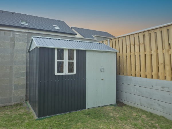 The 10ft x 6ft Steel Cottage shed in a garden. There is a wooden fence to the right of the frame and a large grey wall to the left. The sky is at dusk and there are two houses visible in the backgroun.