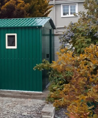 The 8ft x 6ft Steel garden shed in green as seen from the side. A large orange bush is in the forground, to the right of the scene and the shed on the left has a window kit installed in it
