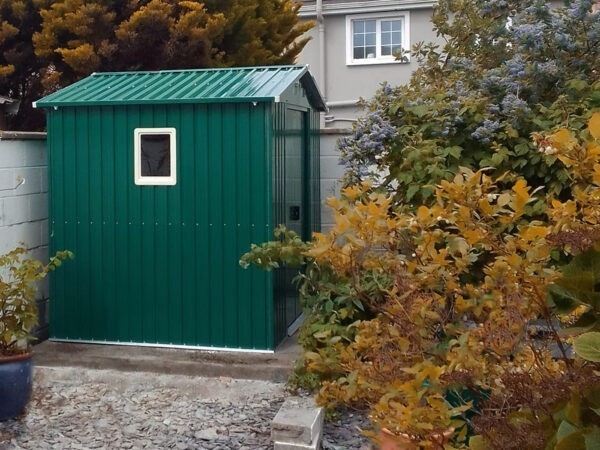 The 8ft x 6ft Steel garden shed in green as seen from the side. A large orange bush is in the forground, to the right of the scene and the shed on the left has a window kit installed in it