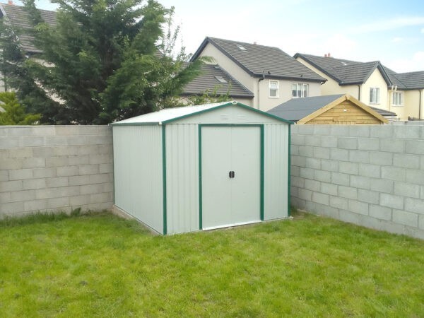 A grey-white 9ft x 10ft steel garden shed in a garden in Limerick. The grass is green and occupies most of the bottom half of the image. The White shed is in a corner, with grey walls surrounding two sides of it. Beyond
