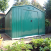 The Green Colossus shed in a garden at sunset with plants all around the shed and a wooden railing behind it