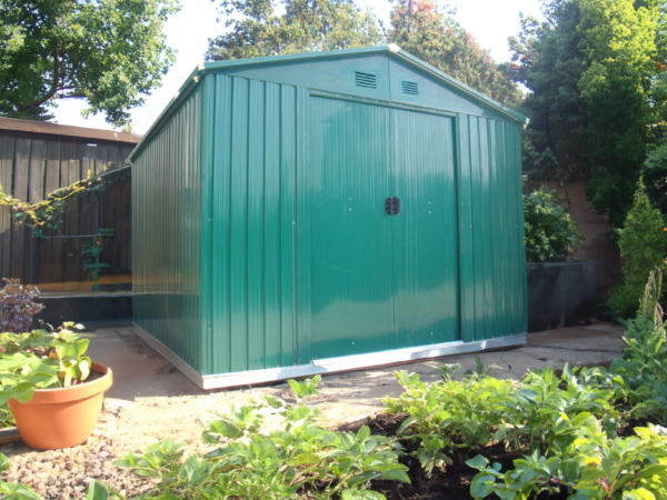 The Green Colossus shed in a garden at sunset with plants all around the shed and a wooden railing behind it