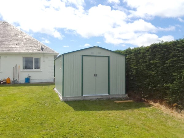 A customer's photo of the 10ft x 12ft colossus shed in white. It's stands on a concrete base on a green, grassy lawn, beside a large bush with a large, bungalow house behind it.