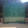 The goldilocks shed against a small brick wall that is only 1 foot high. There are dense bushes above the wall which enclose the green, metal shed on both sides.