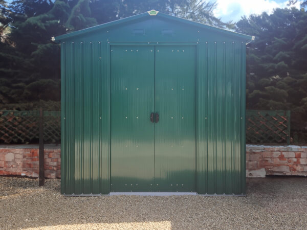 The goldilocks shed against a small brick wall that is only 1 foot high. There are dense bushes above the wall which enclose the green, metal shed on both sides.