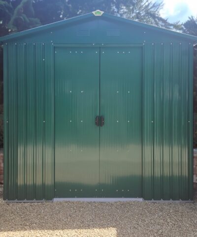 The goldilocks shed against a small brick wall that is only 1 foot high. There are dense bushes above the wall which enclose the green, metal shed on both sides.