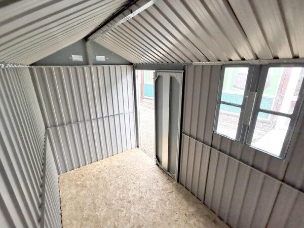 An interior view of the cottage shed from the far end of the shed, looking towards the door. White light pours in from the open doorway and from the window on the right hand side of the fram