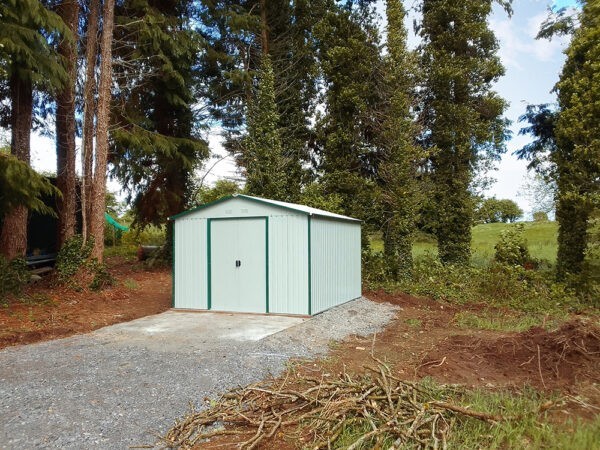 A customer's photo of the off-white Colossus Garden Shed in a large garden with tall, evergreen trees surrounding it.