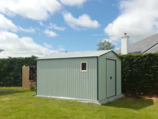 A colossus shed in white as seen from the side. It sits on a solid, level concrete base, which is laid onto grass. There is a large dark green bush behind it and a mostly blue and slightly cloudy sky above. The shed has a window installed in it.