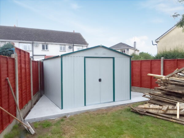 A 10ft x 12ft colossus shed in off-white in a triangular shaped garden. There is a red fence either side of the shed, a large concrete base underneath it and a blue sky above.