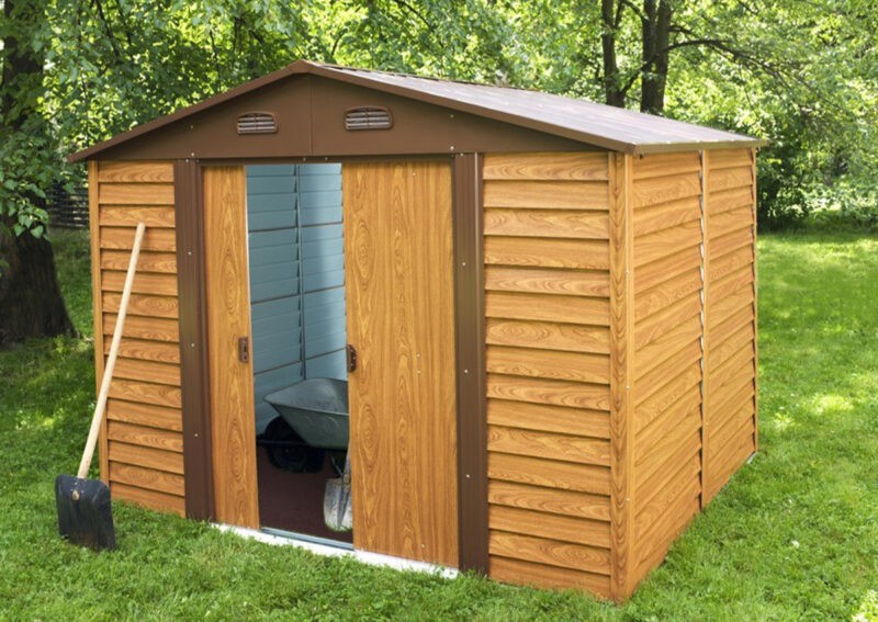 The woodgrain shed seen from a 45 degree angle. The door is open and inside the shed has grey walls and there is a dulled metal wheelbarrow and spade visible. Outside the shed is a bright orange-brown colour and there is grass and trees surrounding it.