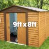 The woodgrain shed seen from a 45 degree angle. The door is open and inside the shed has grey walls and there is a dulled metal wheelbarrow and spade visible. Outside the shed is a bright orange-brown colour and there is grass and trees surrounding it. On top the text reads '9ft x 8ft'