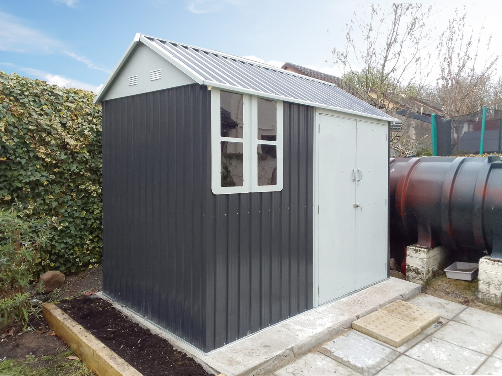 The 8ft x 6ft steel cottage shed assembled in a garden, There is a concrete base beneath it, some slabs leading up to it and an empty flower bed to the side.