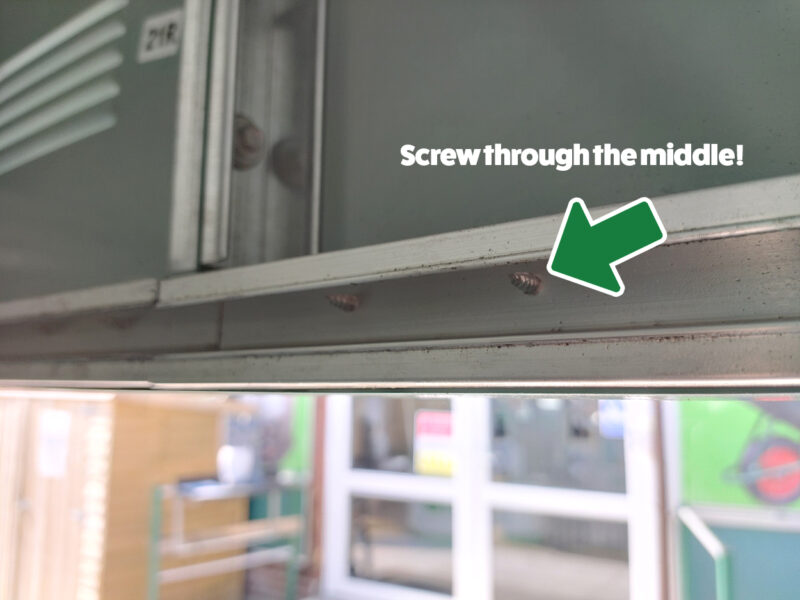 A picture of the inside of a shed, looking at the runner panel above the door, which has a large dip in the middle, between two bars. A green arrow points to the middle and text above reads 'screw through the middle!'