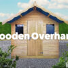 A front view of the Wooden Overhang Shed from Sheds Direct Ireland. This golden-wooden shed has a large, 2 foot protruding roof )i.e. the overhang) which casts a shadow over the top of the shed. There are two windows and a single door on the front of the shed.