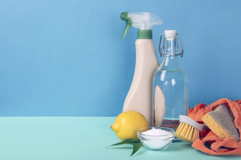 A picture of a lemon, a clear bowl of baking soda, a brush, a jar of vinegar and a squirt-cap cleaning bottle against a tow tone blue background