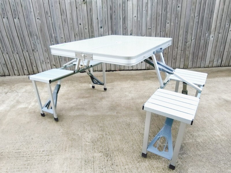 A wide angled view of the picnic table. It is almost silver in colour and the table top is a very pale grey. There is a wooden wall in the background.