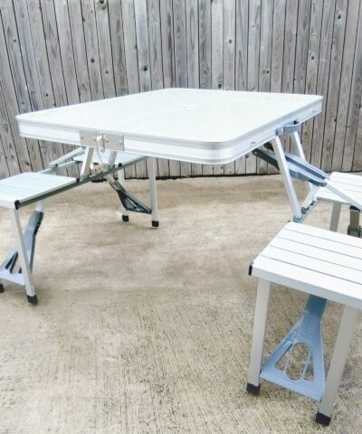 A wide angled view of the picnic table. It is almost silver in colour and the table top is a very pale grey. There is a wooden wall in the background.