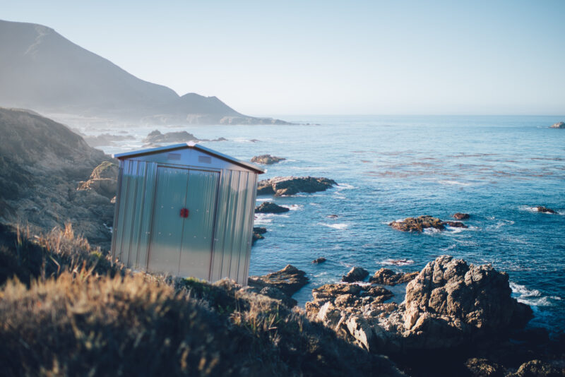 A shed photoshopped into a rocky field overlooking the coast. The shed is at a comically jaunty angle and the rest of the scene is very idyllic, with foamy white eaves, a cool blue sky and rugged pale-green hills 