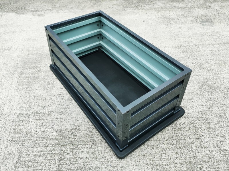 An above view of a raised metal garden bed. It's an anthracite colour on the outside and a paler grey on the inside. The base is a black-rubber looking material. It stands on a grey concrete base. 