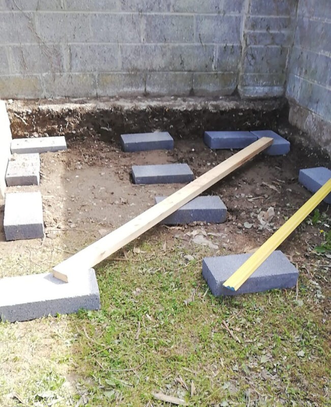 A base for a shed comprised of 10 grey concrete blocks spaced relatively well with two different planks of wood placed horizontally across them