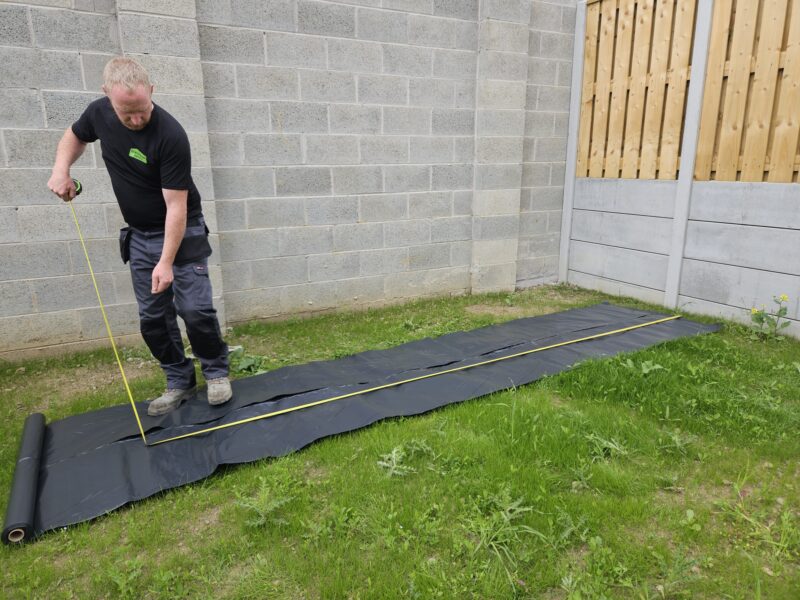 A man in a black t shirt and working trousers measuring a roll of damp-proof membrane using a bright yellow tape. The membrane sits on top of the green lawn and the area is walled in by grey stone. 