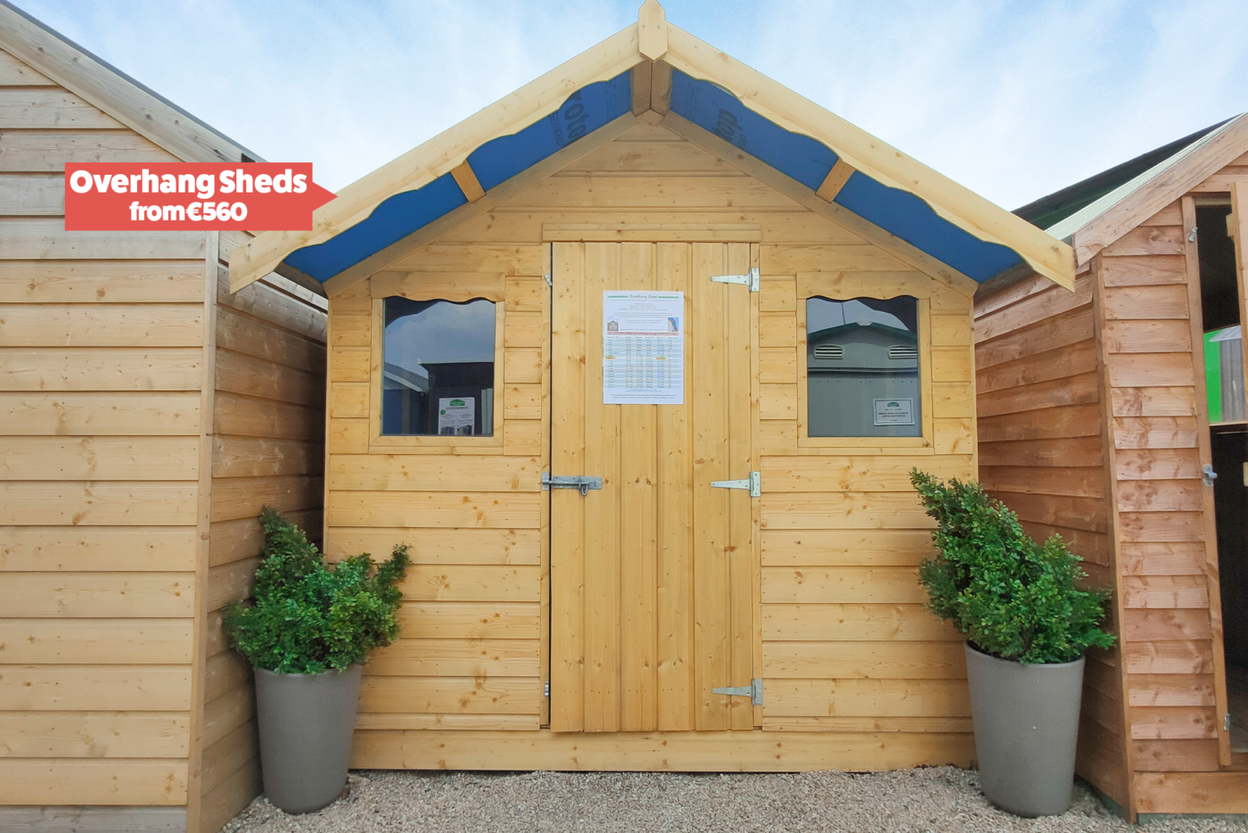 A picture of a wooden overhang shed with two dark windows and two plant pots either side of the door.