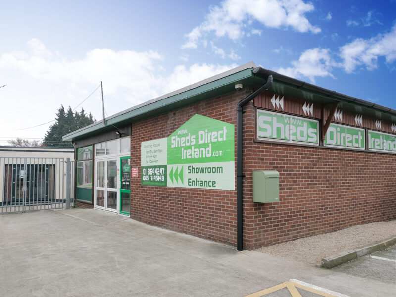 The exterior of the Sheds Direct Ireland showroom. It's a redbrick building with green vinyl-stickers of the Sheds Direct Ireland logo along it. There is a grey railing to the side of the entrance and a pale-green post box on the side of the building. 