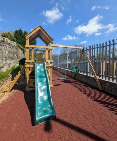 The Alpha+ Playhouse on a red-brick base with stone wall to the left. There is a green slide and green swings
